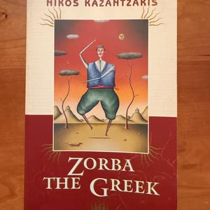 May include: A paperback book titled Zorba the Greek by Nikos Kazantzakis. The cover features a surreal illustration of a person dancing in a landscape with a red sky, green trousers, and a blue vest, set against a cream and deep red background.