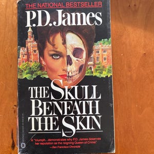 Puede incluir: Una portada de libro de bolsillo con fondo negro y un rostro de mujer dividido a la mitad con una calavera. El título del libro es "The Skull Beneath The Skin" de P.D. James. El texto de la portada dice "A "triumph...demonstrates why P.D. James deserves her reputation as the reigning Queen of Crime." -San Francisco Chronicle"
