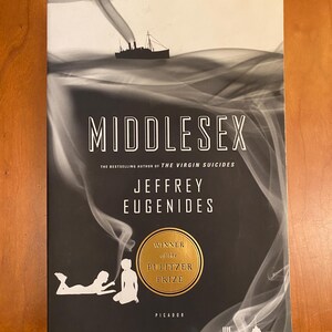 May include: A hardcover book titled "Middlesex" by Jeffrey Eugenides. The cover features a black and white design with a ship, smoke, and silhouettes. It has a gold seal that says "Winner of the Pulitzer Prize".
