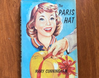 Może przedstawiać: Okładka starej książki z kolorową ilustracją kobiety w kapeluszu. Tytuł książki to "The Paris Hat" autorstwa Mary Cunningham.