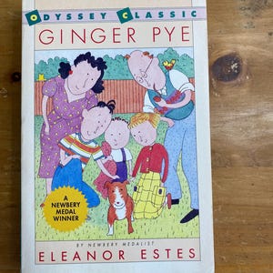 Può includere: Una copia vintage di "Ginger Pye" di Eleanor Estes. La copertina del libro presenta un'illustrazione colorata di una famiglia con un cane. Il titolo è in lettere rosse e il libro è etichettato come vincitore della medaglia Newbery.