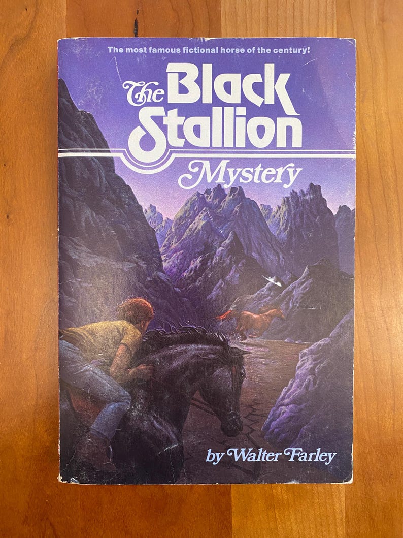May include: The Black Stallion Mystery book cover features a black horse galloping through a purple mountain range with a man riding on its back. The text on the cover reads "The Black Stallion Mystery" and "by Walter Farley".
