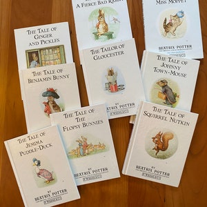 May include: A collection of Beatrix Potter children's books, featuring titles like "The Tale of Peter Rabbit" and "The Tale of Jemima Puddle-Duck." The books have white covers with illustrations of animals and text. The books are arranged on a wooden surface.