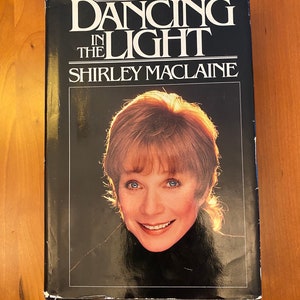 May include: A black hardcover book with the title "Dancing in the Light" by Shirley Maclaine. The cover features a photo of Shirley Maclaine smiling.