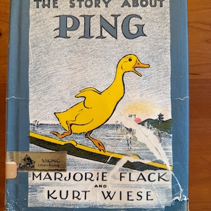 May include: A hardcover book titled The Story About Ping by Marjorie Flack and Kurt Wiese. The blue cover features a yellow duck standing on a boat, with a landscape illustration in the background. A Viking Library Binding sticker is visible.