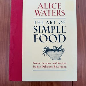 Può includere: Un libro di cucina intitolato "ALICE WATERS THE ART OF SIMPLE FOOD" con dorso rosso. La copertina presenta testo e un'illustrazione di un cestino. Il sottotitolo recita "Notes, Lessons, and Recipes from a Delicious Revolution."