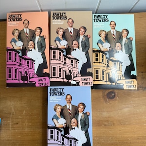 May include: A collection of five VHS tapes of the British sitcom "Fawlty Towers." Each tape features the show's title and a cast photo. The tapes have different colored backgrounds, including peach, tan, and blue.
