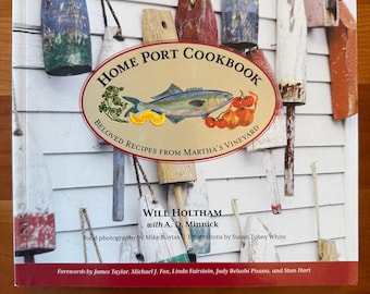 May include: A cookbook titled "Home Port Cookbook" with the subtitle "Beloved Recipes from Martha's Vineyard." The cover features an illustration of a fish, tomatoes, and lemons. The background shows weathered wooden planks and buoys.