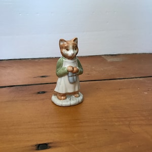 以下が含まれることがあります： 緑色のエプロンを着て、小さく青と白のパッケージを持った猫のセラミック製フィギュア。猫はオレンジと白の毛皮で、白い台座の上に立っています。