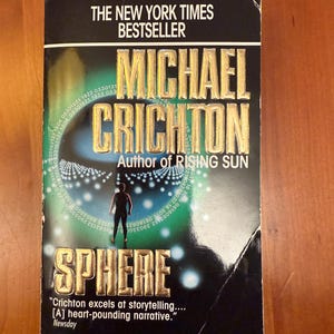 May include: A paperback book titled "Sphere" by Michael Crichton. The cover features a silhouetted figure, a blue and white design, and the text "The New York Times Bestseller". The book is a Ballantine/Fiction publication.