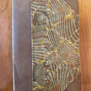 Pu&ograve; includere: Un libro rilegato in pelle marrone con una copertina marmorizzata in tonalit&agrave; di verde, marrone e oro. La copertina presenta un motivo decorativo di linee e forme vorticose.