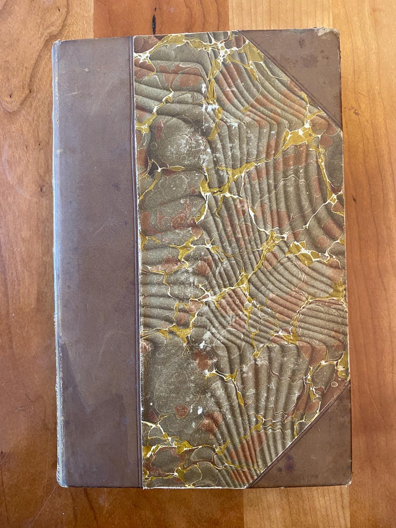 Pu&ograve; includere: Un libro rilegato in pelle marrone con una copertina marmorizzata in tonalit&agrave; di verde, marrone e oro. La copertina presenta un motivo decorativo di linee e forme vorticose.