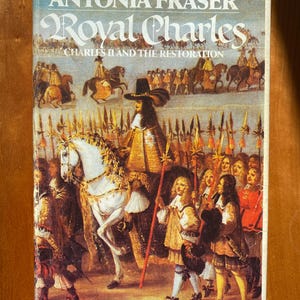 Op de afbeelding: Een boekomslag met een witte achtergrond en de titel "Royal Charles" in zwarte cursieve letters. De ondertitel is "Charles II en de Restauratie". De omslag toont een schilderij van Charles II te paard, omringd door een groep mensen.