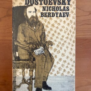 May include: Vintage book cover featuring a seated illustration of a man with a long beard. The title reads "DOSTOEVSKY NICHOLAS BERDYAEV" in bold black letters. The background is a cream color with a floral pattern. Published by Meridian Books.