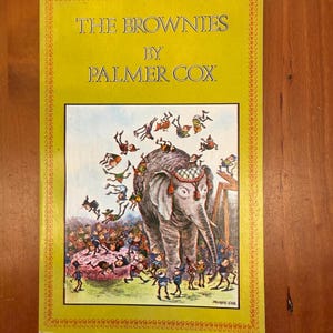 May include: A vintage book titled "The Brownies by Palmer Cox" with a yellow cover and a detailed illustration. The illustration depicts an elephant surrounded by small figures in colorful outfits, with text in a serif font.