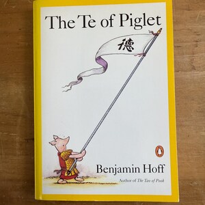 Puede incluir: Un libro titulado "The Te of Piglet" de Benjamin Hoff. La portada presenta a Piglet sosteniendo una bandera con caracteres chinos. El libro tiene un borde amarillo y un logotipo de Penguin. El autor es también de "The Tao of Pooh".