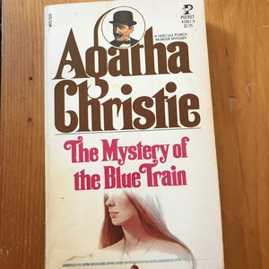 以下が含まれることがあります： 長い金髪の女性が茶色と白で描かれた、ペーパーバックの表紙。本のタイトルは「青い列車の謎」アガサ・クリスティ著。表紙には帽子をかぶった男性の小さなイラストも描かれている。
