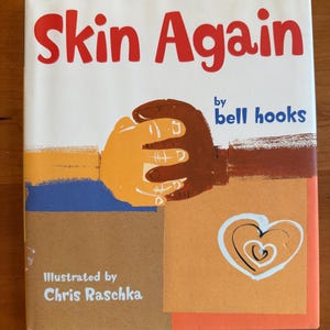 Könnte beinhalten: Das Buch Skin Again von bell hooks, illustriert von Chris Raschka, zeigt ein Cover mit rotem Text und einer Illustration von zwei ineinander verschränkten Händen unterschiedlicher Hauttöne. Ein weißes Herz ist auf beigem Grund.