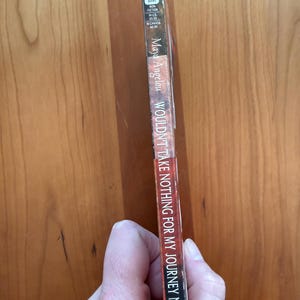 K&ouml;nnte beinhalten: Ein Taschenbuch von Maya Angelou mit dem Titel "Wouldn't Take Nothing For My Journey Now". Der Buchr&uuml;cken ist schwarz mit wei&szlig;em Text und einem rot-wei&szlig;en Logo. Das Buch wird vor einem Holzgrund gehalten.