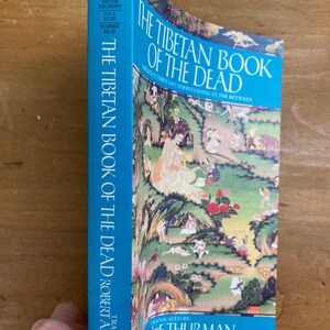 Puede incluir: Un libro azul titulado "El Libro Tibetano de los Muertos: Liberaci&oacute;n a trav&eacute;s de la comprensi&oacute;n del intermedio" de Robert A.F. Thurman, traducido por el Dalai Lama. La portada presenta una ilustraci&oacute;n colorida de un paisaje con figuras.