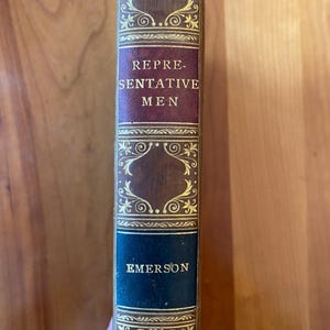 Pu&ograve; includere: Un libro vintage con una copertina in pelle marrone e lettere dorate. Il titolo &egrave; "Representative Men" di Emerson.