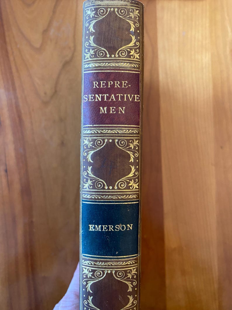 Pu&ograve; includere: Un libro vintage con una copertina in pelle marrone e lettere dorate. Il titolo &egrave; "Representative Men" di Emerson.