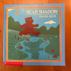 May include: A children's book cover titled "Bear Shadow" by Frank Asch. The cover features a brown bear jumping over a stream, with its reflection in the water. The background includes trees, flowers, and a blue sky.