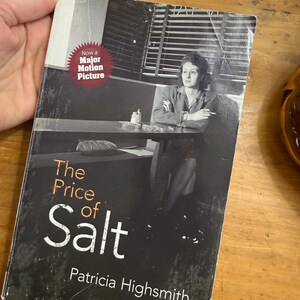 May include: A paperback book titled The Price of Salt by Patricia Highsmith. The black and white cover features a person sitting at a table with a cigarette. A red circular badge on the cover reads Now a Major Motion Picture in white text.