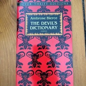 May include: A paperback book titled "THE DEVIL'S DICTIONARY" by Ambrose Bierce. The cover is red with a repeating black devil face pattern. The book is part of the Dover Thrift Editions series.