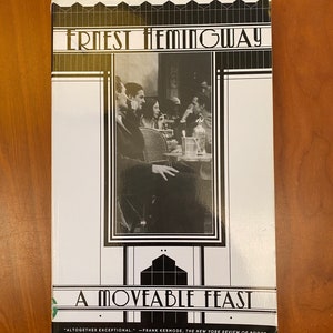 May include: A paperback book titled A Moveable Feast by Ernest Hemingway. The cover features a black and white photograph of people at a cafe table, framed by black and white geometric lines. The text reads: Ernest Hemingway, A Moveable Feast.