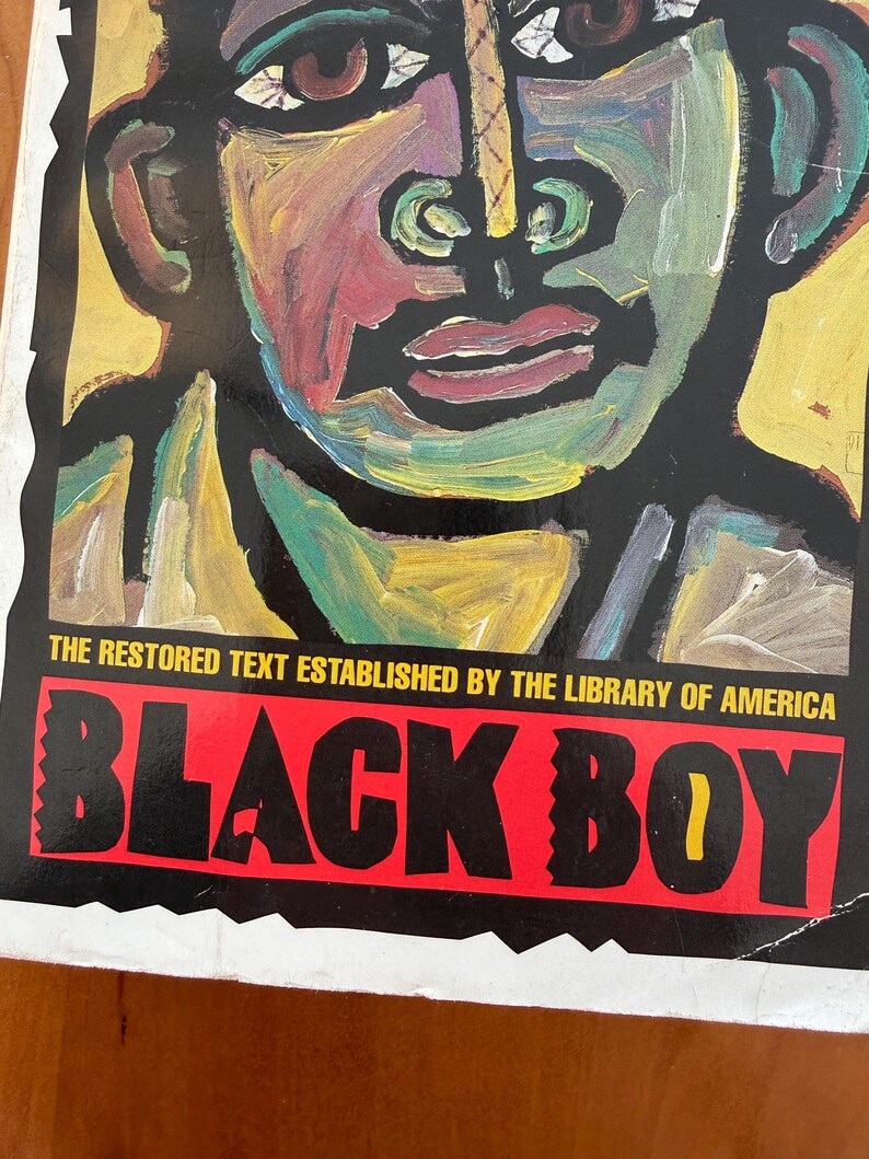 Op de afbeelding: Een boekomslag met een kleurrijk, abstract portret tegen een zwarte achtergrond. De titel "BLACK BOY" staat in vette, witte letters op een rode banner. De tekst "THE RESTORED TEXT ESTABLISHED BY THE LIBRARY OF AMERICA" staat boven de titel.