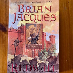 Könnte beinhalten: Ein Taschenbuch mit dem Titel "Redwall" von Brian Jacques. Das Cover zeigt eine Burgszene mit einem Mäusekrieger, der ein Schwert und einen Schild hält. Der Titel ist in großen roten Buchstaben gehalten, mit dem Text "The Legend Begins!" darunter.