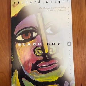 May include: A paperback book titled Black Boy by Richard Wright. The cover features a colorful, abstract painted portrait of a face in shades of yellow, purple, red, and black. The text reads The Restored Text Established by The Library of America.
