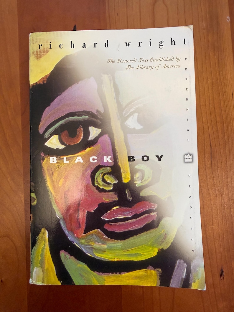 May include: A paperback book titled Black Boy by Richard Wright. The cover features a colorful, abstract painted portrait of a face in shades of yellow, purple, red, and black. The text reads The Restored Text Established by The Library of America.