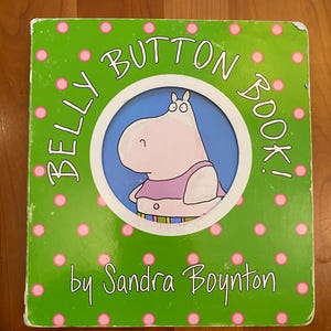 Op de afbeelding: Een kinderboek getiteld "BELLY BUTTON BOOK!" door Sandra Boynton. De cover is groen met roze stippen en toont een cartoon nijlpaard in een cirkel. Het nijlpaard draagt een paars shirt en heeft een navel.