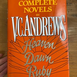 May include: A hardcover book with an orange cover featuring the text "THREE COMPLETE NOVELS BY V.C. ANDREWS" in white and gold lettering. The book's title, "Heaven, Dawn, Ruby," is also displayed in gold. The cover design includes a stylized sun.