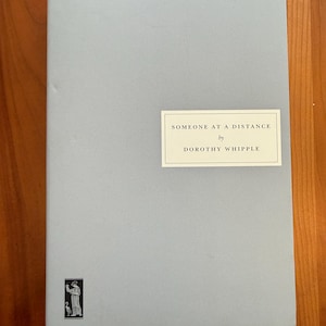 Puede incluir: Un libro azul claro titulado "Someone at a Distance" de Dorothy Whipple. El libro tiene una etiqueta rectangular con el título y el nombre del autor. Una pequeña ilustración en blanco y negro está en la esquina inferior izquierda.