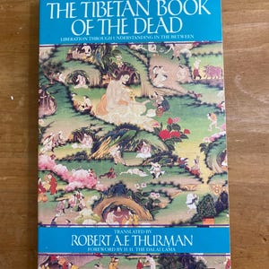 Puede incluir: El Libro Tibetano de los Muertos: Liberaci&oacute;n a trav&eacute;s de la comprensi&oacute;n en el intermedio, traducido por Robert A.F. Thurman con un pr&oacute;logo de S.S. el Dalai Lama. La portada presenta una ilustraci&oacute;n colorida de una escena pac&iacute;fica con muchas figuras.