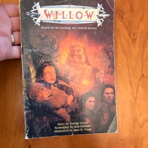 May include: A vintage paperback book titled "WILLOW" based on the motion picture. The cover features a fantasy illustration with characters and the text "Story by George Lucas, Screenplay by Bob Dolman, Adaptation by Joan D. Vinge."