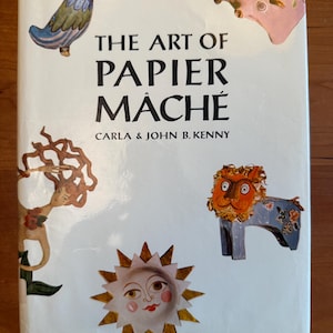 May include: A white hardcover book titled The Art of Papier Mâché by Carla & John B. Kenny. The cover features colorful illustrations of a blue bird, a pink floral head, a stylized figure with long hair, a lion, and a sun with a human face.