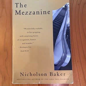以下が含まれることがあります： 黄色い背景に黒い文字で「The Mezzanine」というタイトルが書かれた本の表紙。タイトルの下には、同じく黒い文字で著者の名前「Nicholson Baker」が書かれている。本の表紙には、エスカレーターの白黒写真も掲載されている。