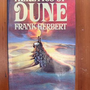 May include: A hardcover book titled "HERETICS OF DUNE" by Frank Herbert. The cover features a surreal landscape with a large, fortress-like structure and a planet in the background. The color palette includes shades of red, orange, and blue.