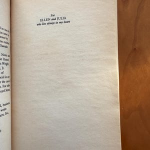 Op de afbeelding: Open boekpagina met de tekst: "For ELLEN and JULIA who live always in my heart." De pagina is gebroken wit met een licht getextureerd oppervlak. De rand van het boek is links zichtbaar en de achtergrond is een warm houten oppervlak.