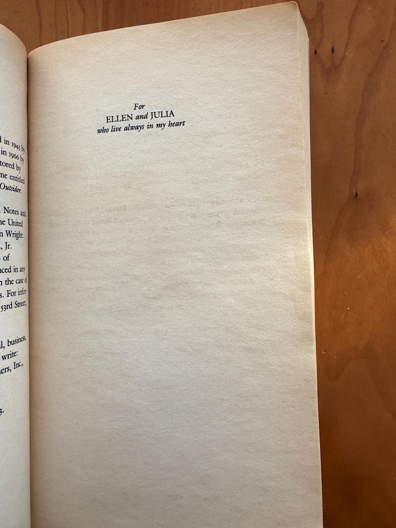 Op de afbeelding: Open boekpagina met de tekst: "For ELLEN and JULIA who live always in my heart." De pagina is gebroken wit met een licht getextureerd oppervlak. De rand van het boek is links zichtbaar en de achtergrond is een warm houten oppervlak.