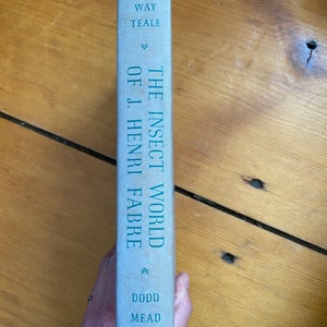 May include: A vintage book titled "The Insect World of J. Henri Fabre" by Edwin Way Teale. The book's spine is light blue with teal lettering. The publisher's name, Dodd Mead, is also visible. The book is held against a wooden surface.