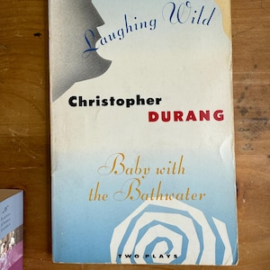 Puede incluir: Una portada de libro vintage con un fondo degradado azul y blanco. El título "Laughing Wild" está en cursiva azul, con el nombre del autor, Christopher DURANG, en negro y rojo. El libro se titula "Baby with the Bathwater".