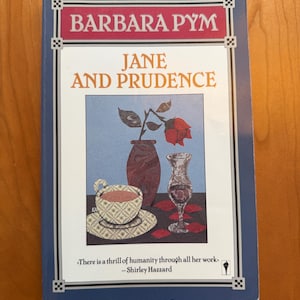 Puede incluir: Libro de Barbara Pym titulado "JANE AND PRUDENCE". La portada presenta un bodegón con una rosa, una taza de té y una copa de vino. Borde azul con el nombre del autor y una cita de Shirley Hazzard.