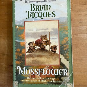 Könnte beinhalten: Ein gebundenes Buch mit dem Titel "Mossflower" von Brian Jacques. Das Cover zeigt eine Illustration von anthropomorphen Mäusen in einer Waldszene. Das Buch gehört zur "New York Times Bestselling Series" und ist ein Prequel zu "Redwall!"