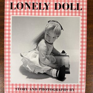May include: The cover of a book titled "THE LONELY DOLL" with a pink and white gingham border. The cover features a black and white photograph of a doll in a dress. The text "STORY AND PHOTOGRAPHS BY DARE WRIGHT" is at the bottom of the cover.