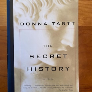 Puede incluir: Una portada de libro con fondo blanco y un primer plano de la cara de una mujer. El título del libro es "The Secret History" de Donna Tartt. El libro se describe como "Una novela" e incluye una cita de The New York Times.